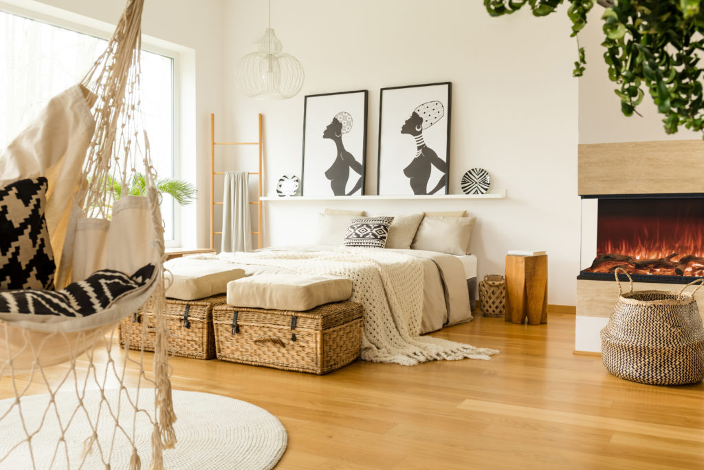 Fireplace near bed with knit blanket in boho bedroom interior with posters and rattan bag